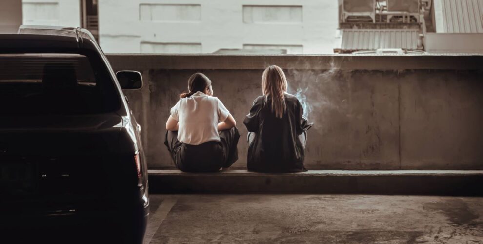 Two Young Women Sitting On Precast Concrete Wheel Stopper And Smokes Cigarette Or Hovering Or Electronic Cigarette At An Empty Parking Lot.